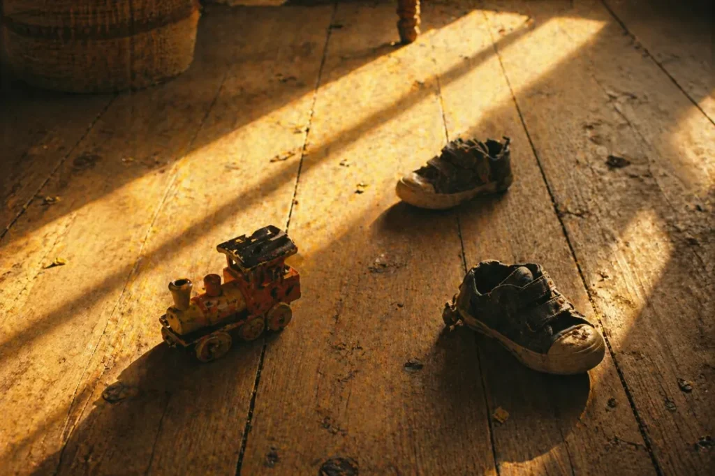 lumière du matin sur un sol en bois avec jouet abîmé et chaussures d’enfant légèrement sales, trace de vie et transmission silencieuse dans la parentalité